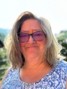 Photo of Diane Coffey standing outside on a sunny day. She has shoulder-length hair and is wearing sunglasses.