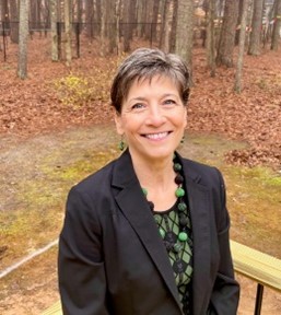 Photo of Virgina Marcus standing outside on a balcony with short brown hair and a black blazer with floral shirt.