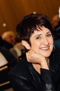 Photo of Holly Riddle sitting at a table with a black blouse on and short black hair, smiling.